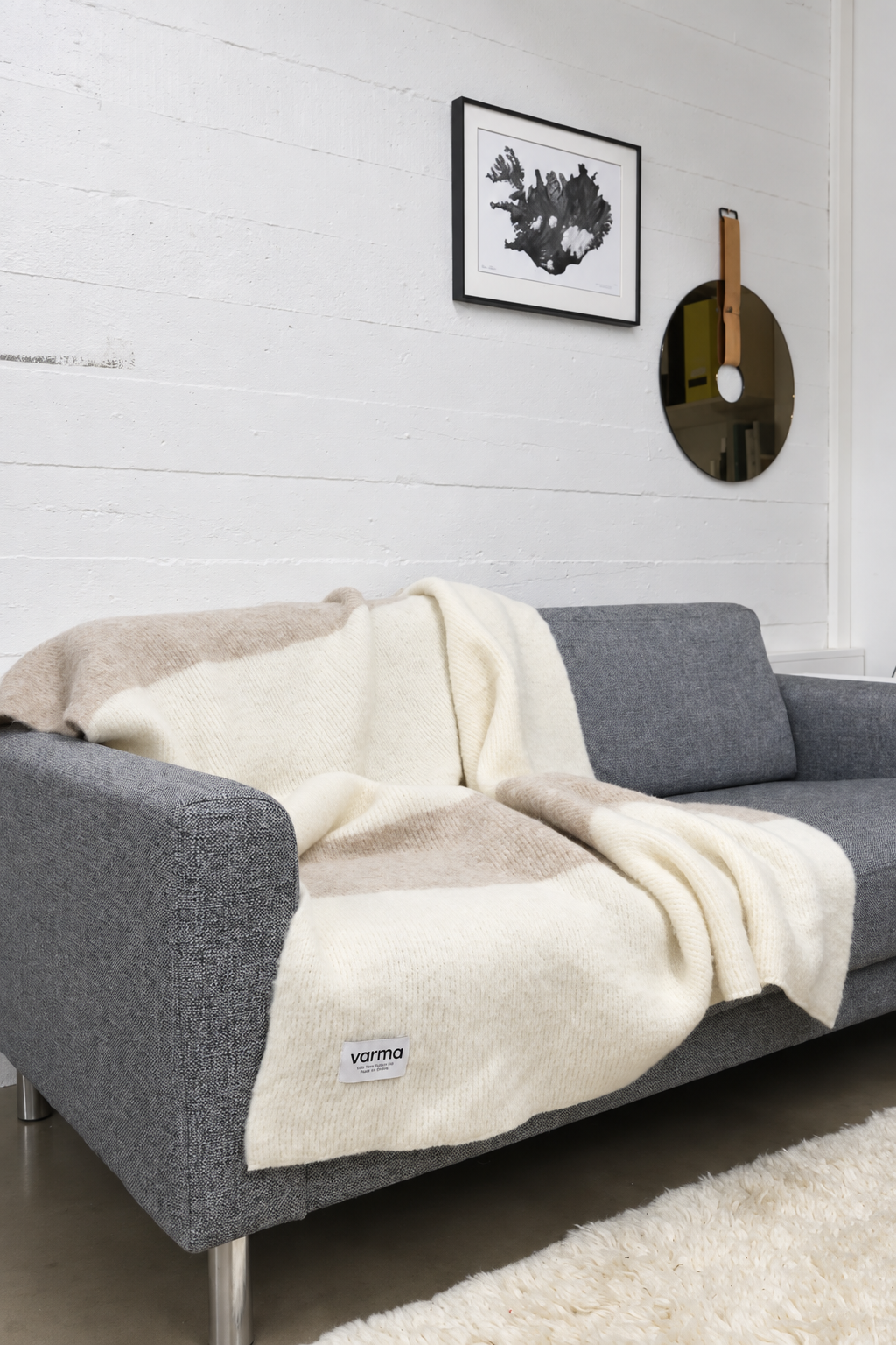A gray fabric sofa in a modern living room setting, draped with a cream and beige striped knit throw blanket. The blanket features a small white woven label near the bottom corner that reads 'varmo'.